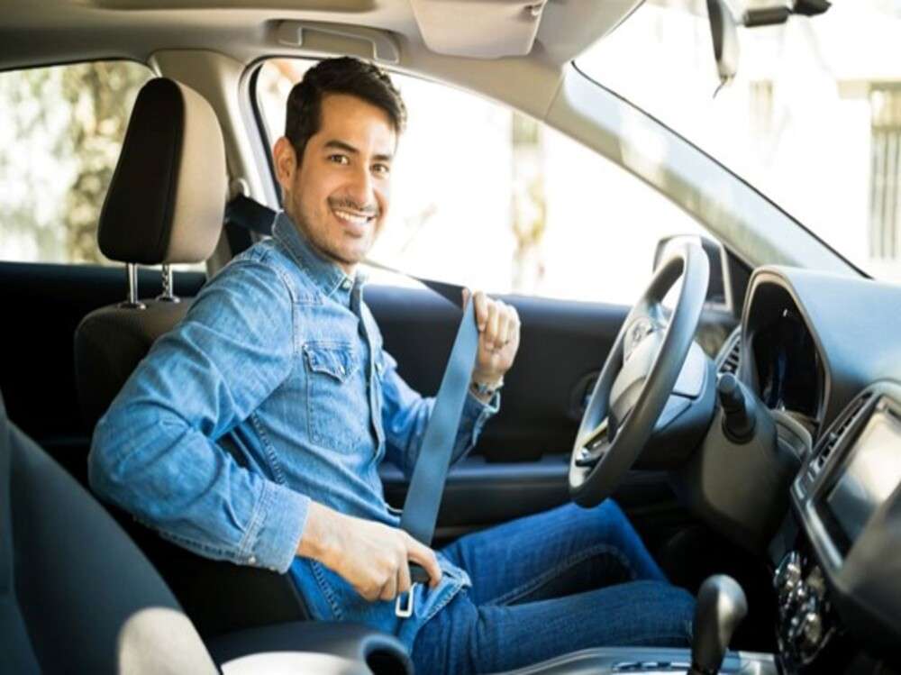 Man in blue shirt in car putting on seat belt smiling.