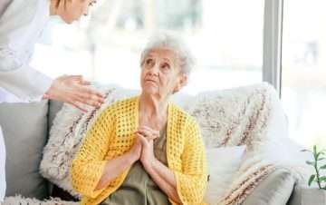 Elderly woman with hands together looking up at woman talking to her.