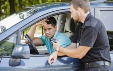 Man in blue shirt in driver seat with hand on head looking concerned. Male police officer at the window talking to him. What are common traffic ticket myths misconceptions that drivers fall for?