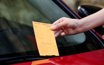 Hand gasping a traffic ticket on the windshield of red car.
